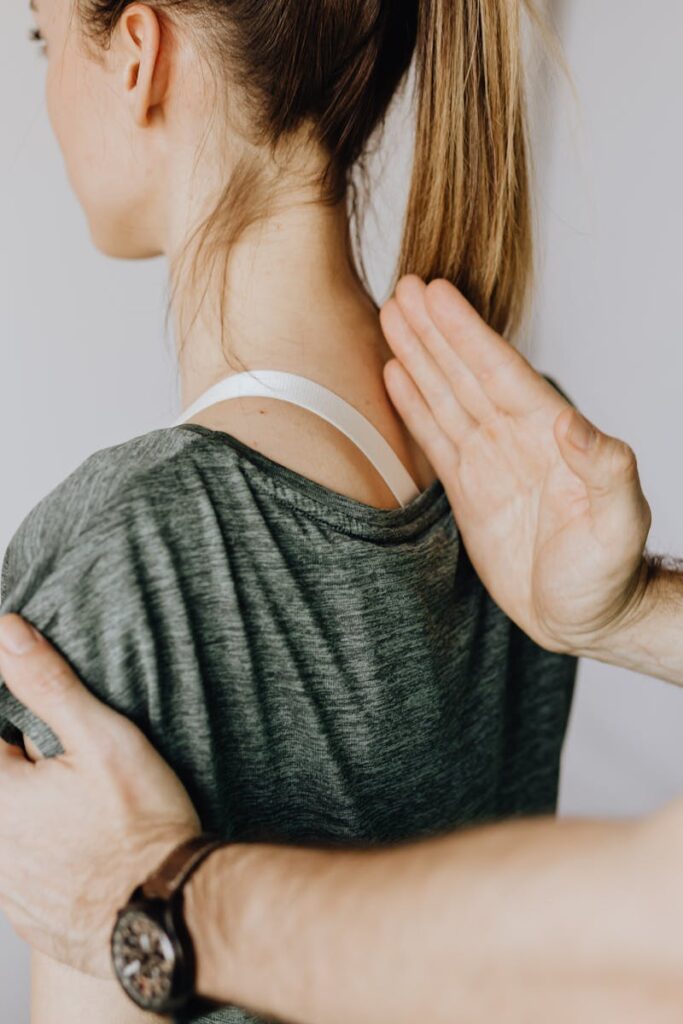 Back view of crop unrecognizable osteopath in uniform and wristwatch checking up back of slim female patient in casual wear on white background